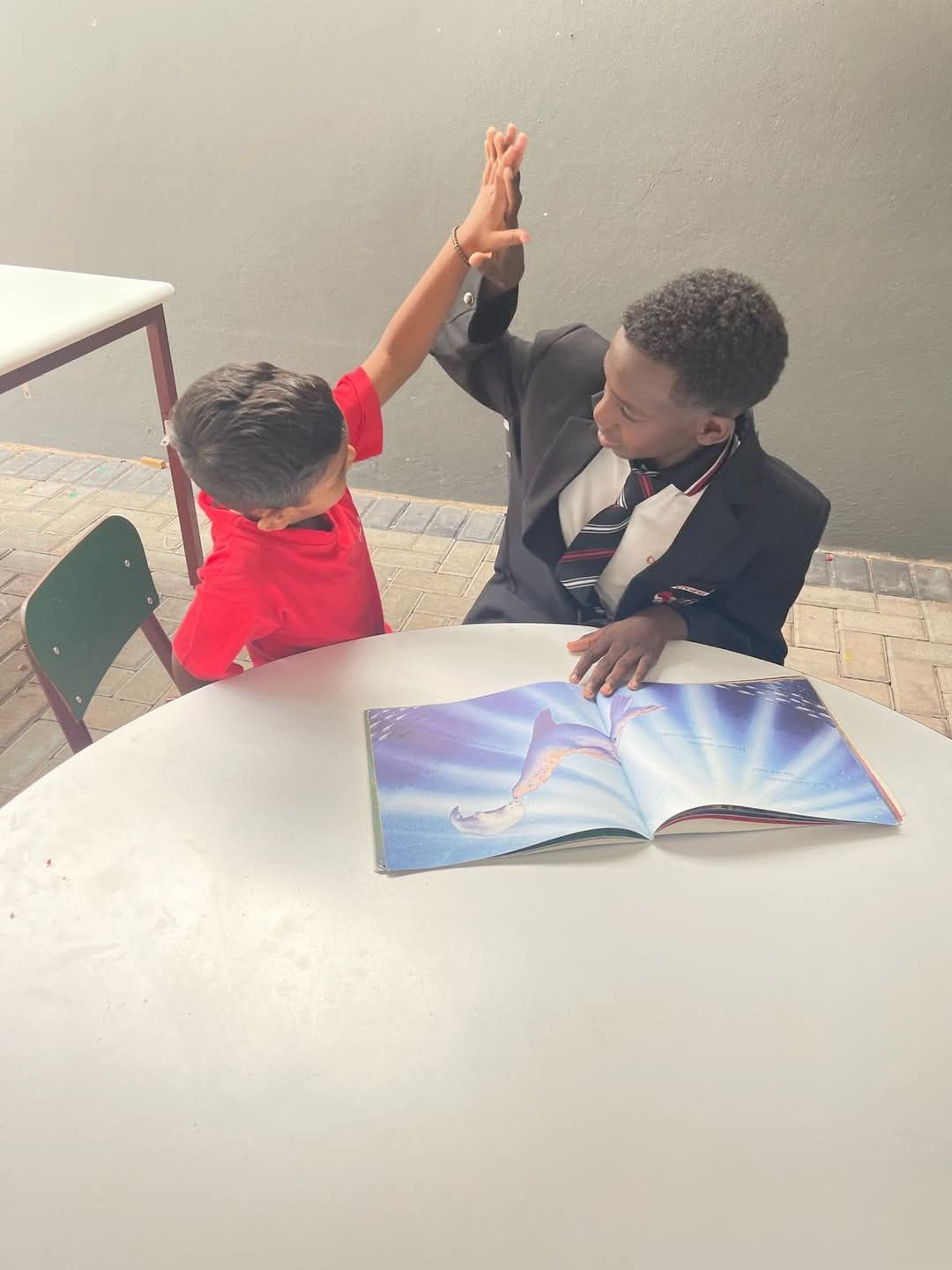 Two children high-fiving over a book at a table; one in a blazer.