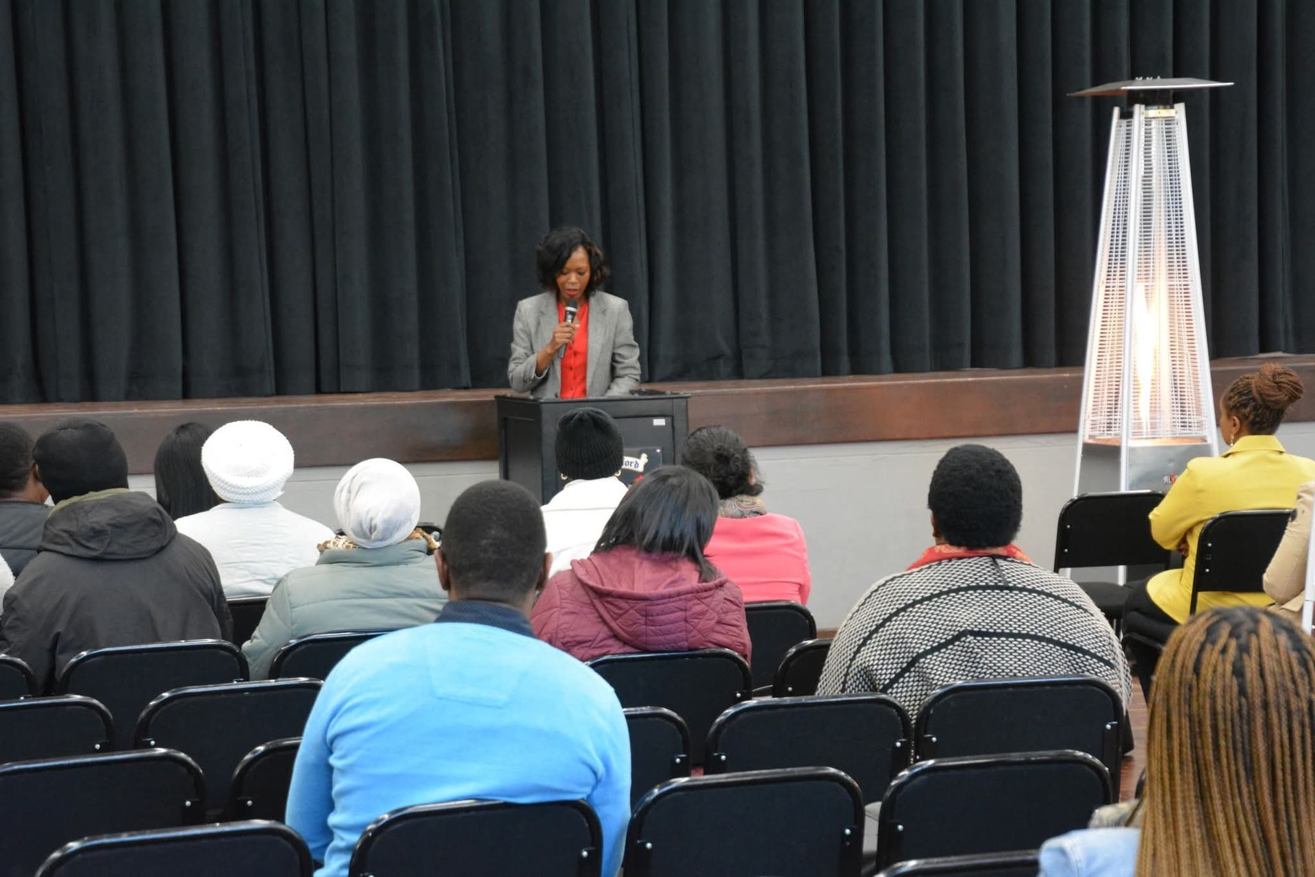 A woman speaks at a podium to an audience in a dark room; a tall heater is on the right.