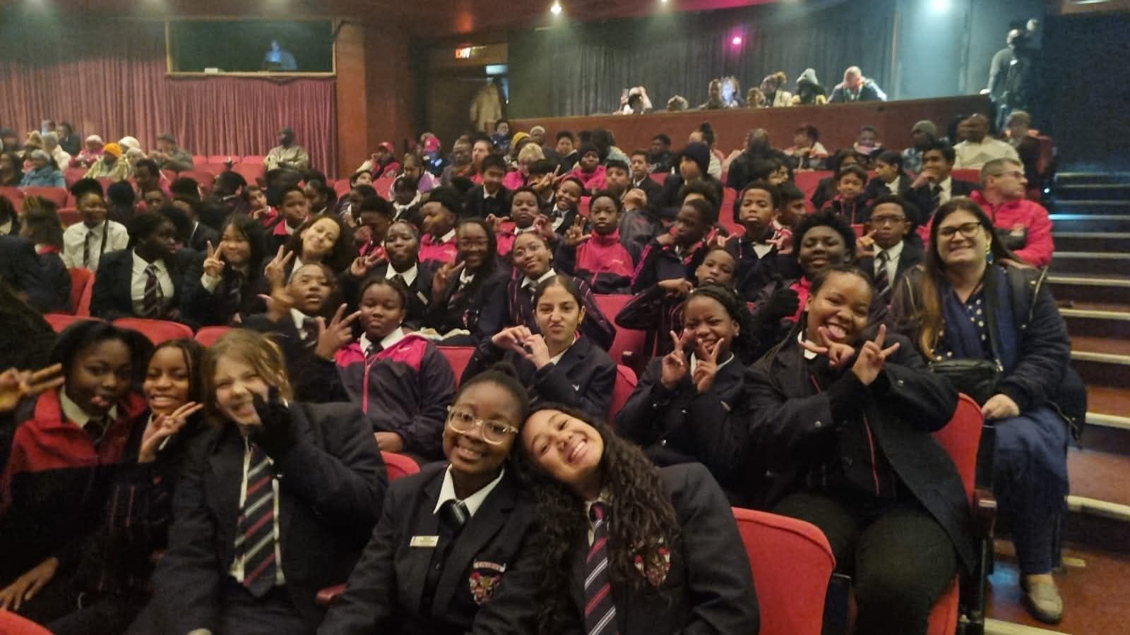 Students in uniform pose in an auditorium, many with peace signs, smiling.