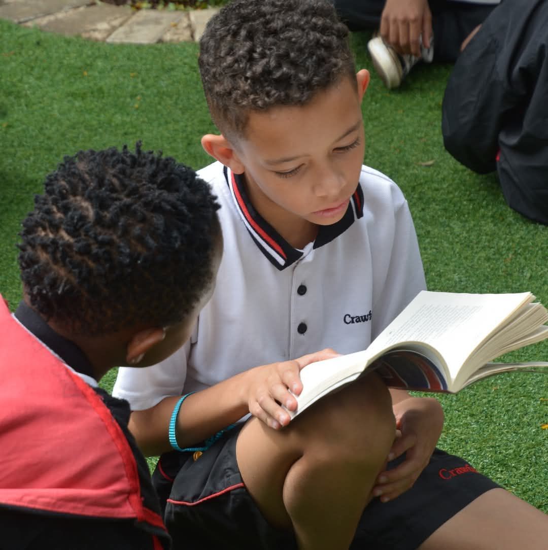 Two students reading a book together on grass; one wears a white collared shirt, the other a red vest.