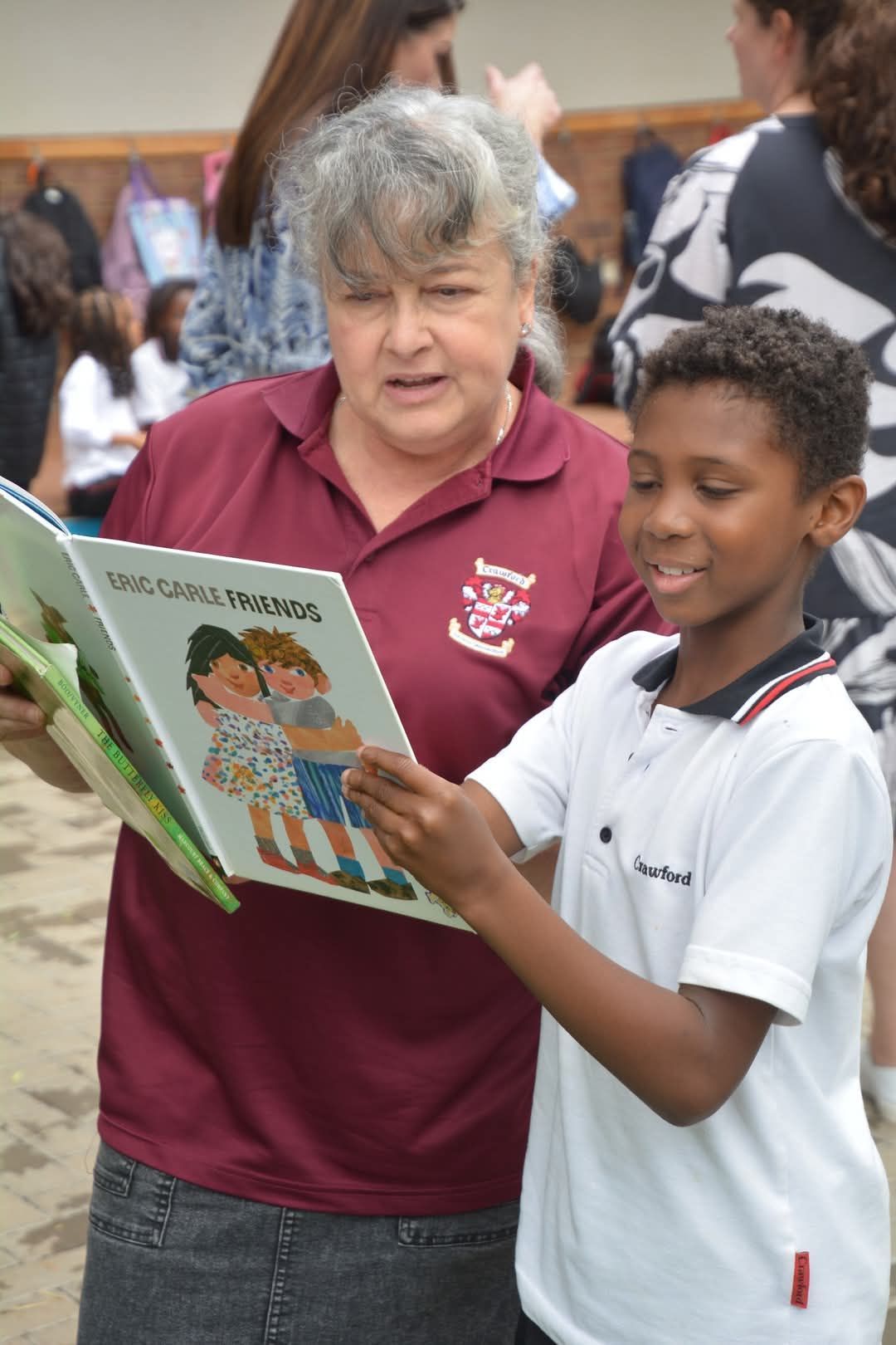 Woman in maroon shirt reads a book with a smiling boy wearing a white polo shirt.