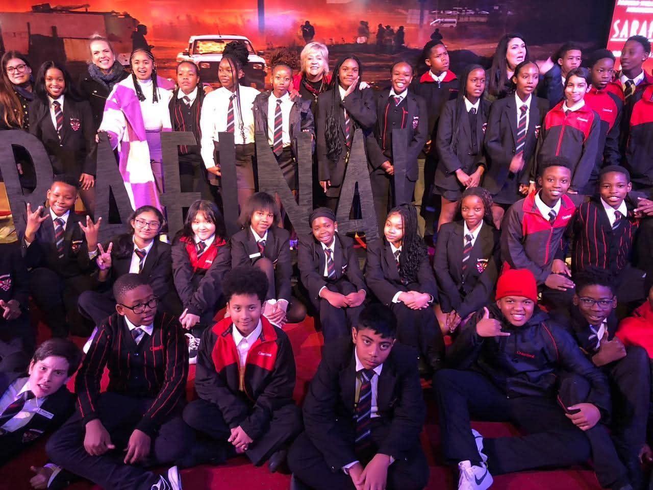 Group of students in school uniforms pose with a sign and background. They are on a red carpet.