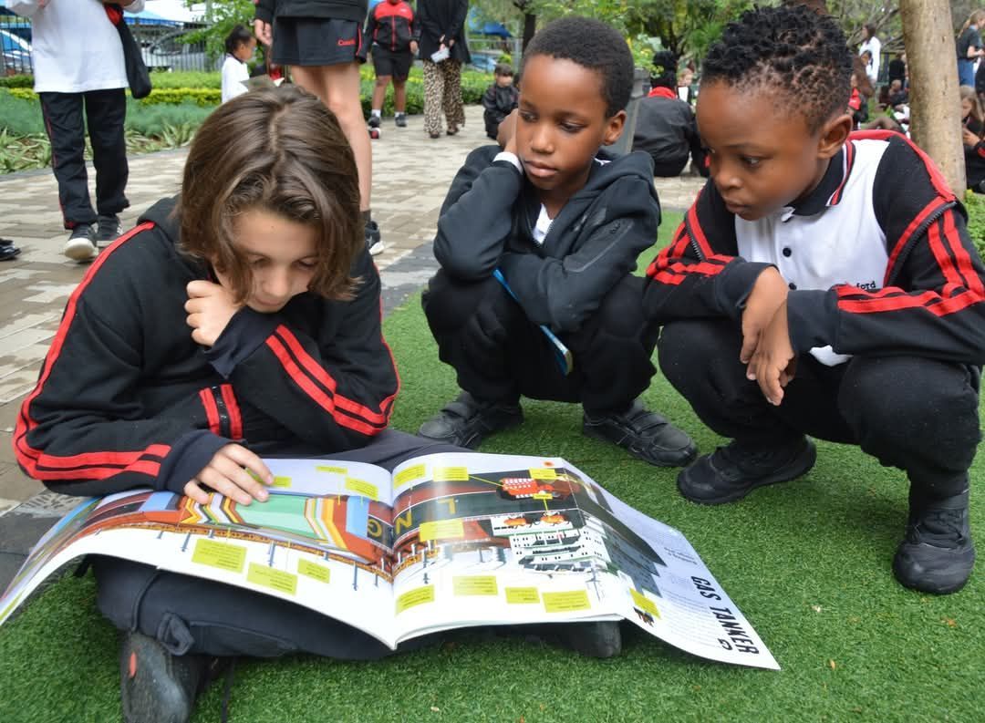 Three students huddled together, reading a book outdoors on artificial turf. All wearing black and red tracksuits.