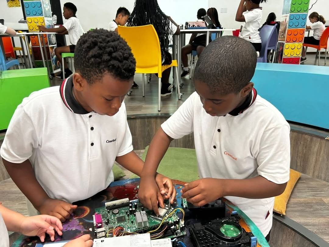 Two children, in white shirts, examine a computer circuit board at a table in a classroom.