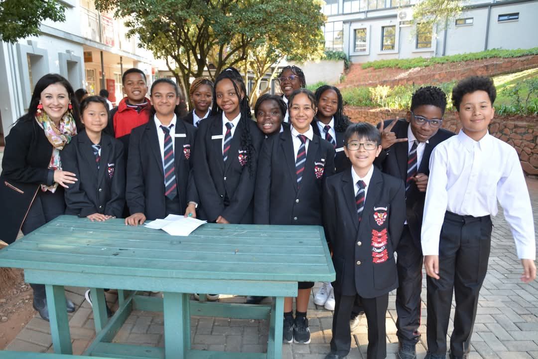Group of students in school uniforms and a woman standing around a green table outside a building.