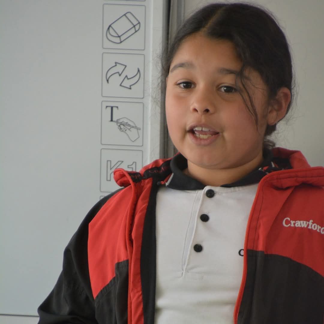 Girl wearing a red and black jacket speaking in front of a whiteboard.