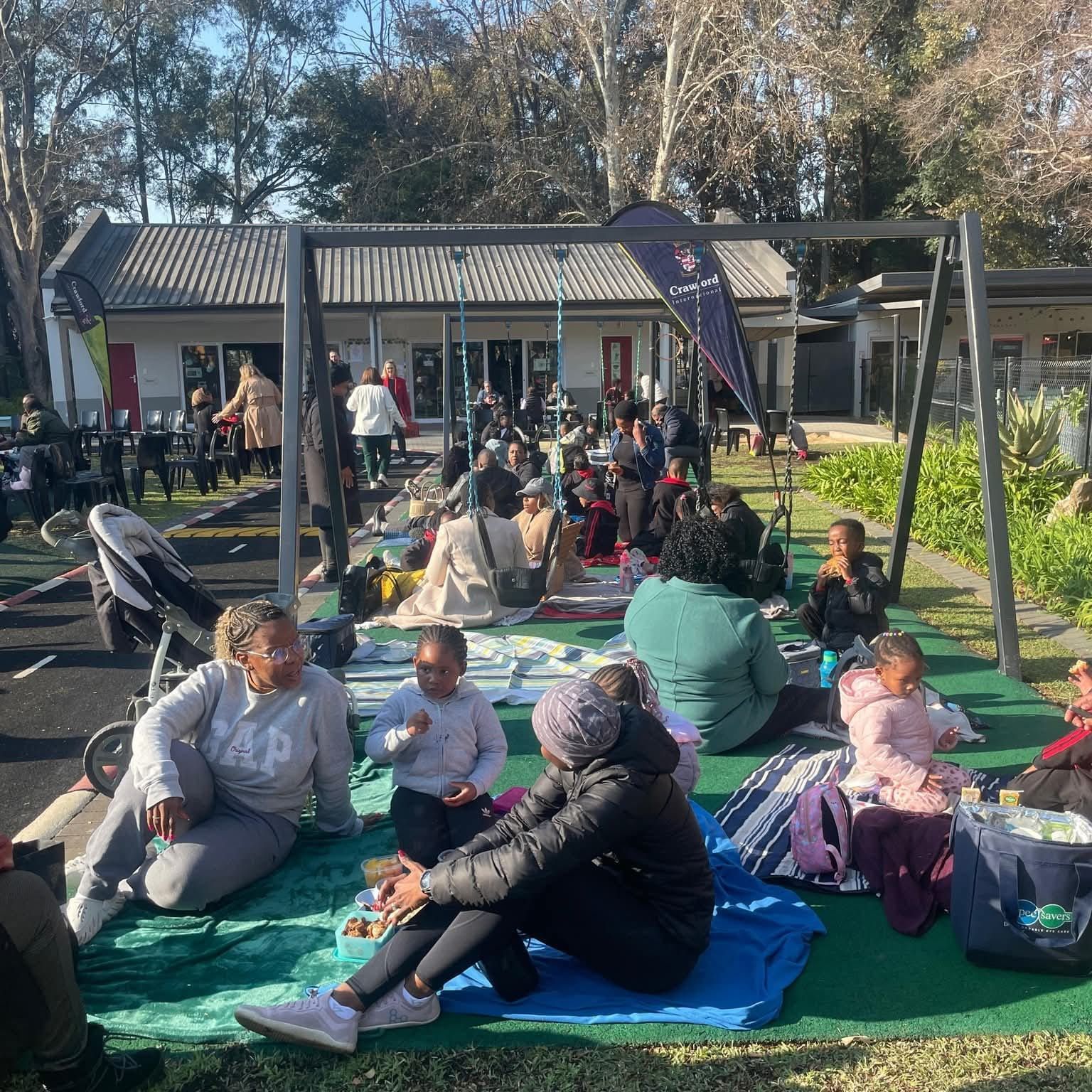 People gathered outdoors on a sunny day. Some are sitting on blankets, and a swing set is visible.