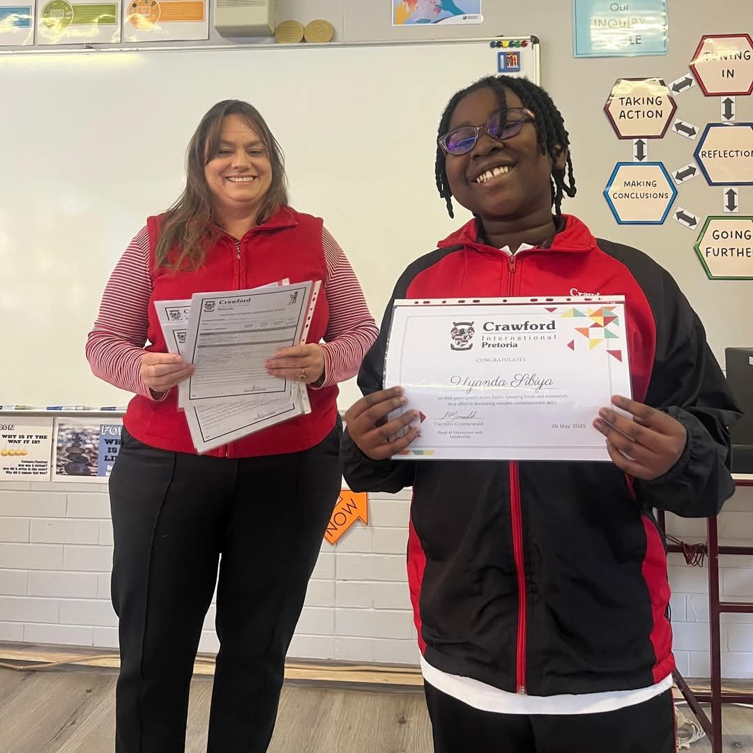 A person holds a certificate, smiling, next to another person holding papers in a classroom.
