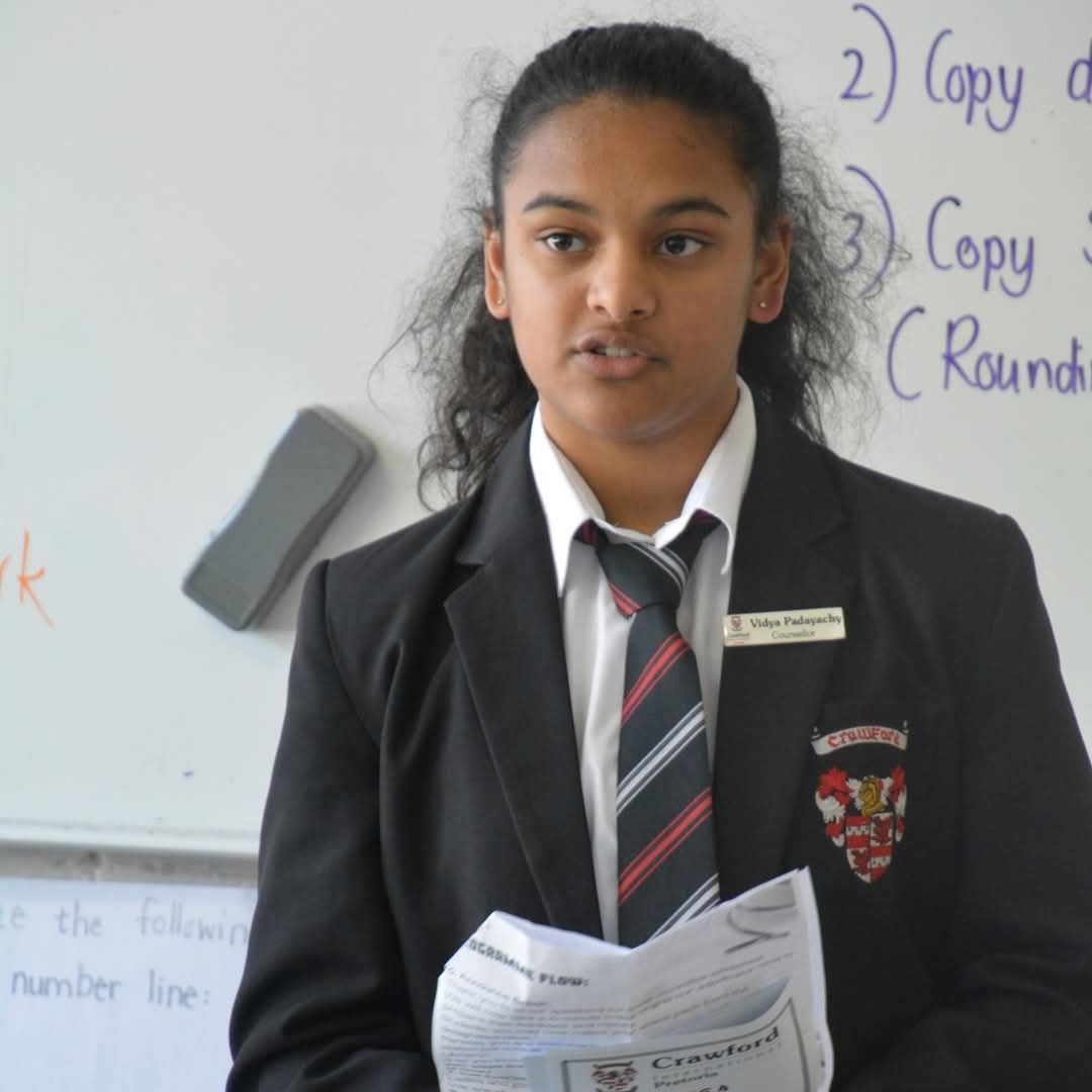 Girl in school uniform giving a presentation, holding papers, near a whiteboard.