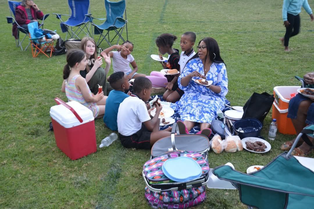 Group of people sitting on grass, eating food, with coolers and chairs nearby.