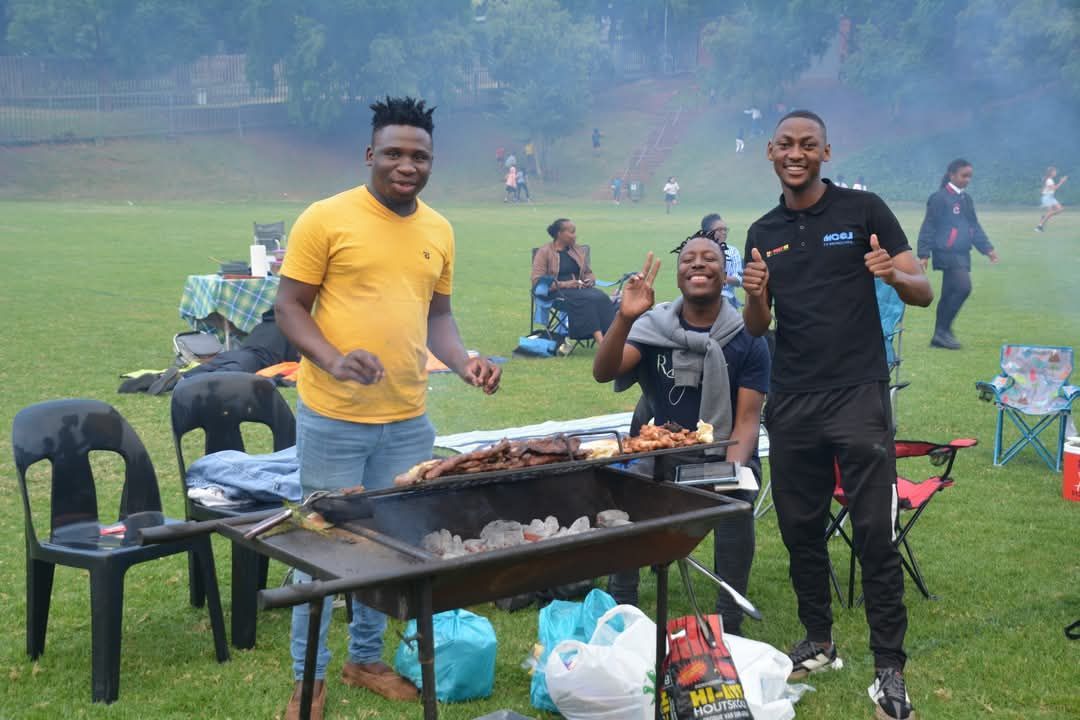 Three people smile by a barbecue grill on a grassy field; grilling food.