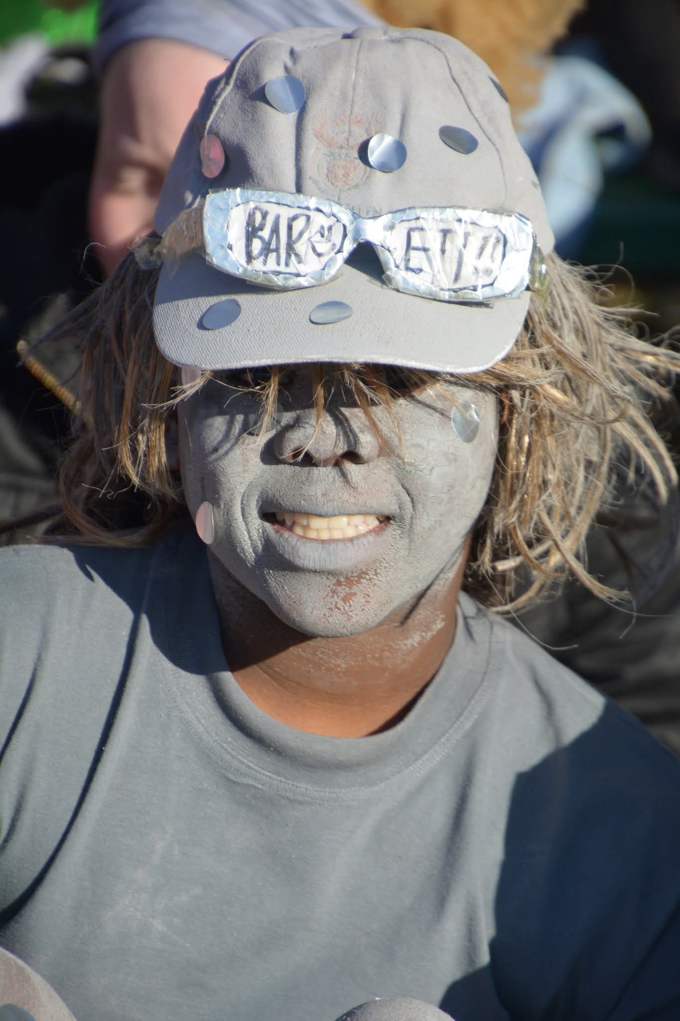Person covered in grey substance, wearing a hat with painted-on sunglasses.