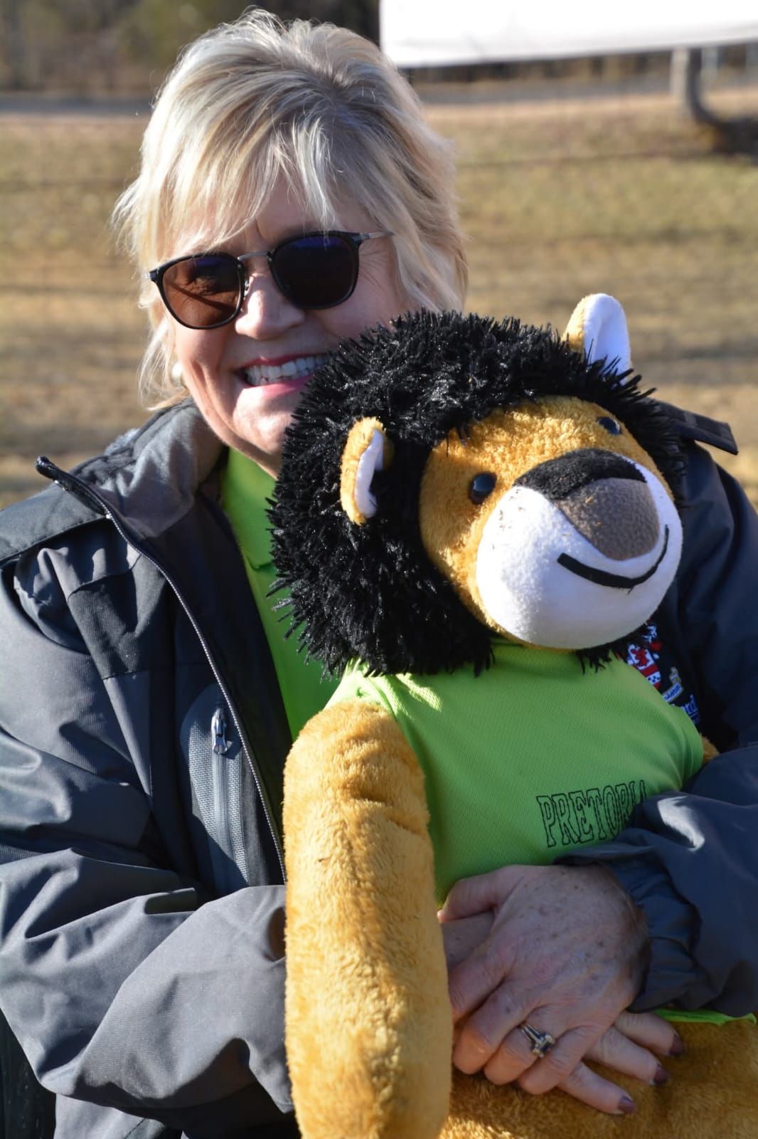 Woman in sunglasses smiles, hugging a stuffed lion wearing a green shirt.