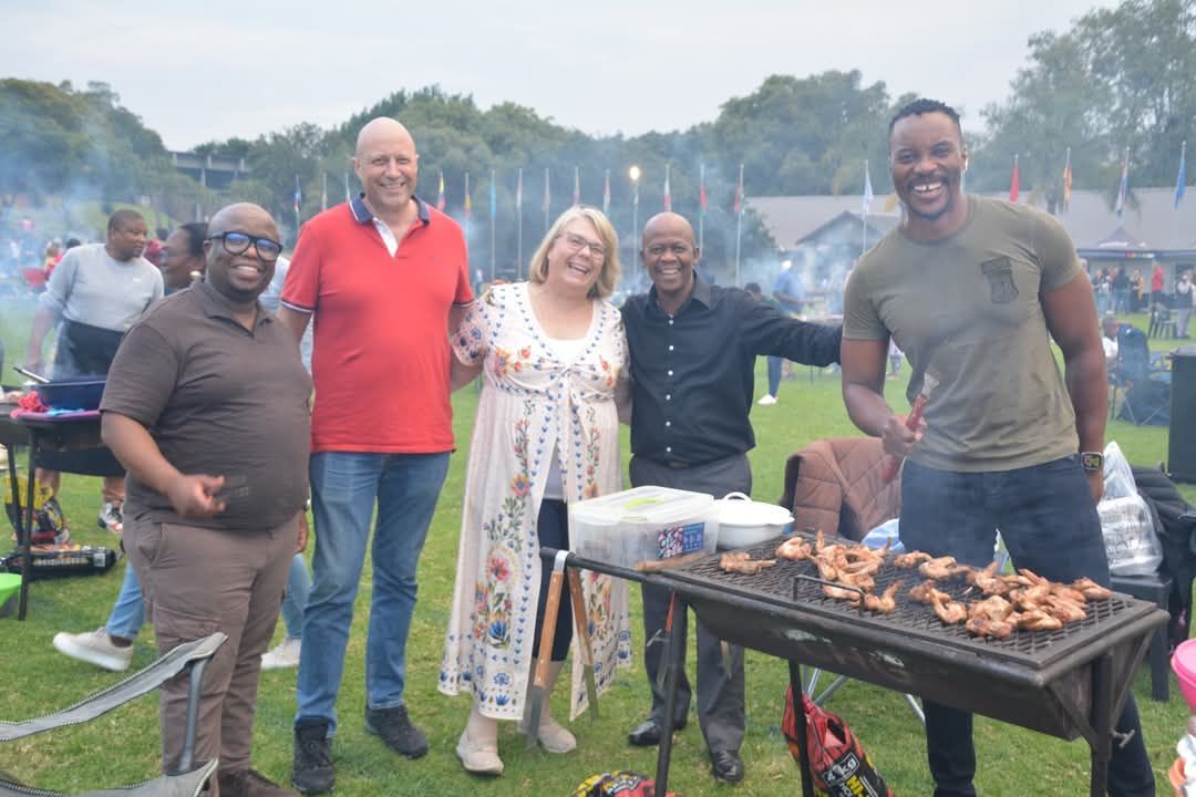 Five people pose near a barbecue grill outdoors, with food cooking.