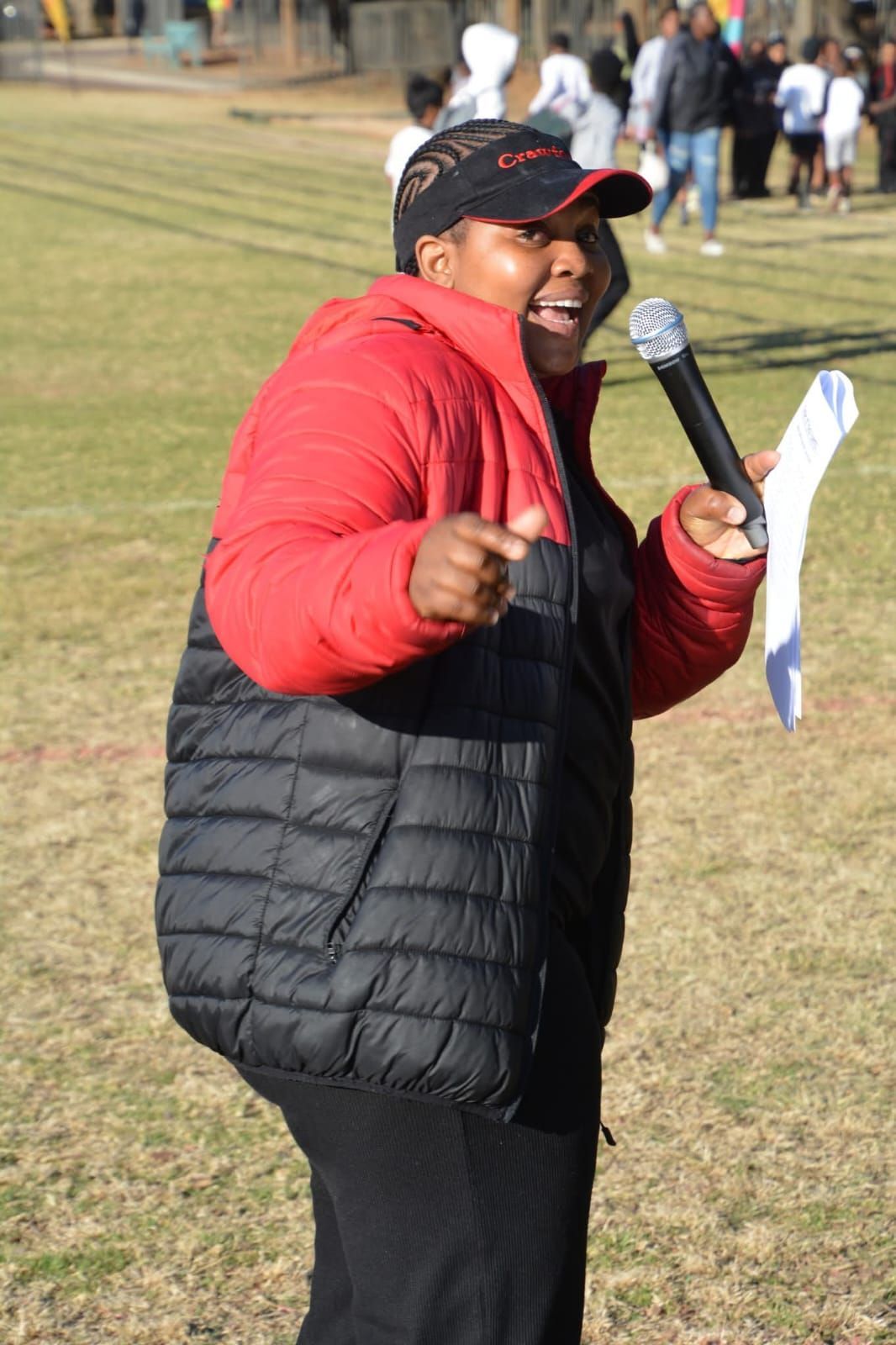 Person in red and black jacket speaking into a microphone at an outdoor event, pointing forward.