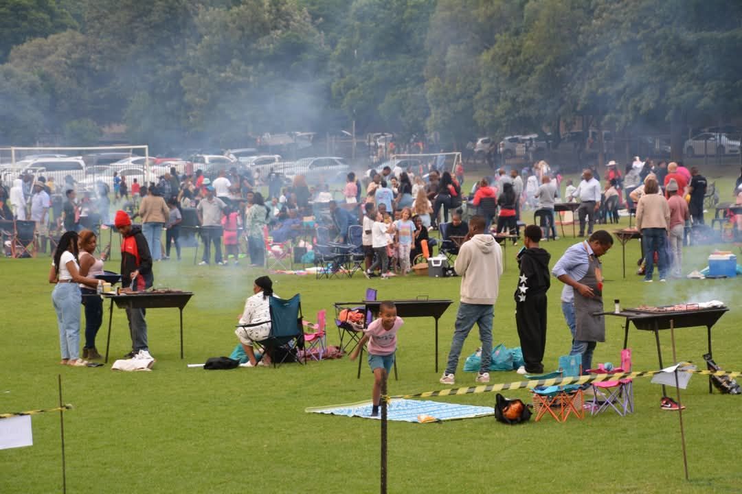 People gathered on a grassy field, grilling food. Smoke rises, cars parked in background.