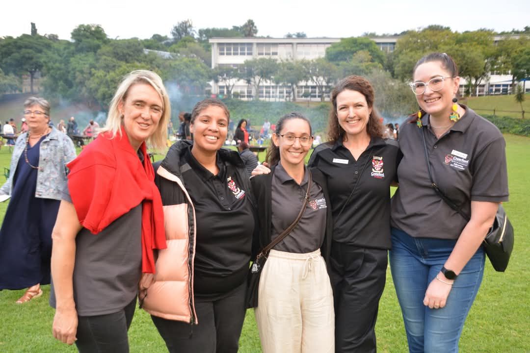 Five people smiling outdoors. Green field, building in background. Two wear black shirts with logo.