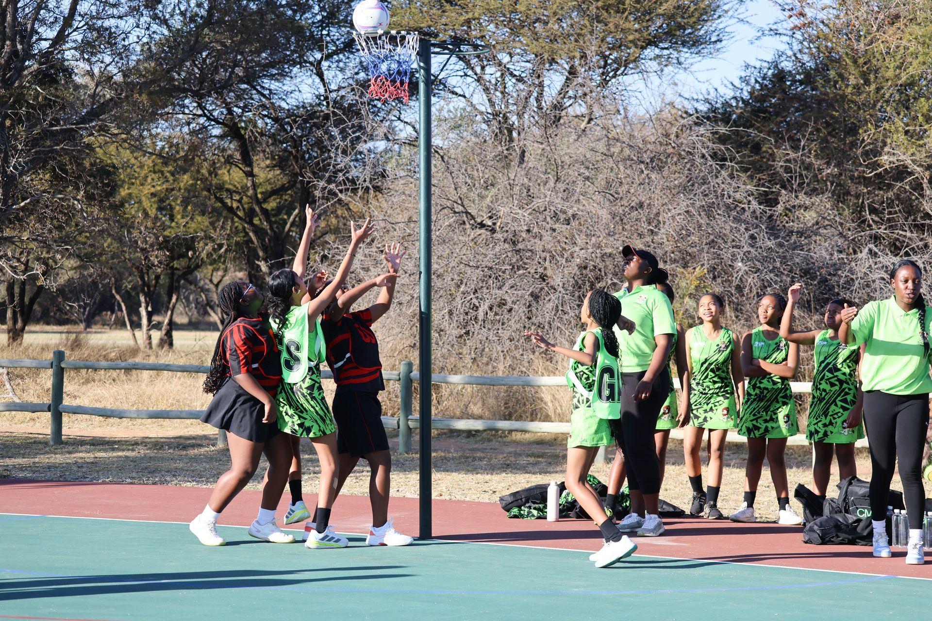 Netball game in progress; players in green and red uniforms jumping to reach the ball near the hoop, outdoors.