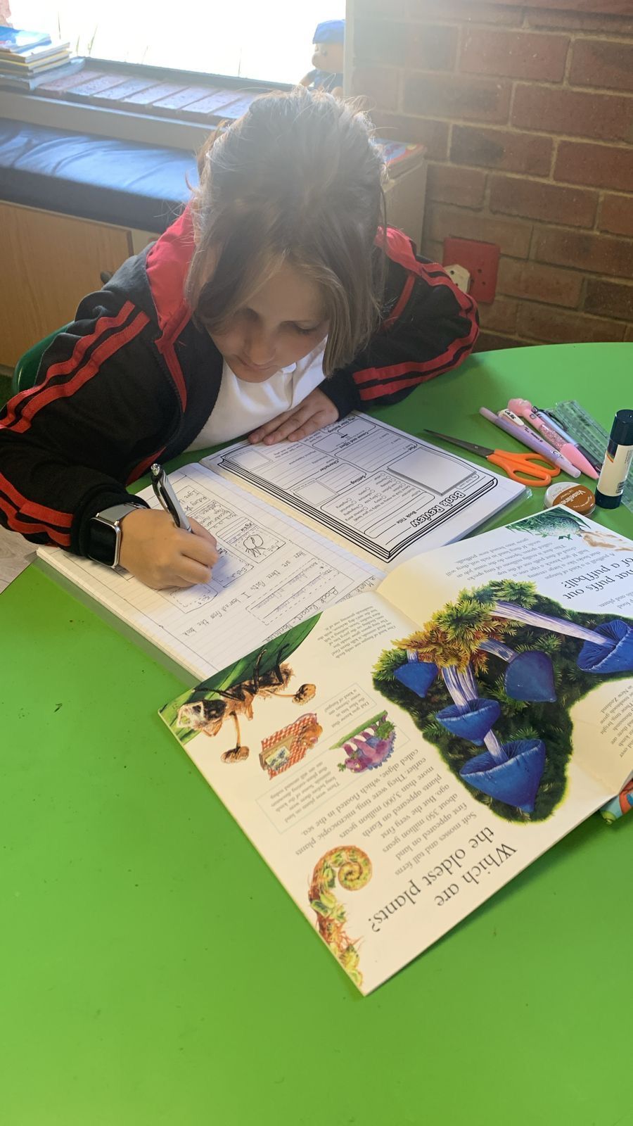 A young girl is sitting at a green table writing in a notebook.