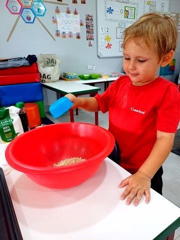 Boy in red shirt pouring from a blue cup into a red bowl with grains. Indoors, bright light.