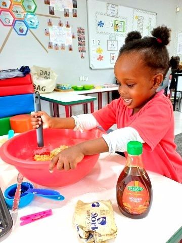 Young child in a red shirt stirring batter in a pink bowl on a table; syrup and butter nearby.