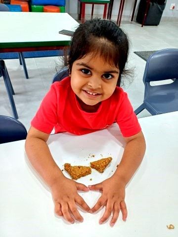 Smiling child in red shirt, hands forming a heart over two pieces of food on a white table.