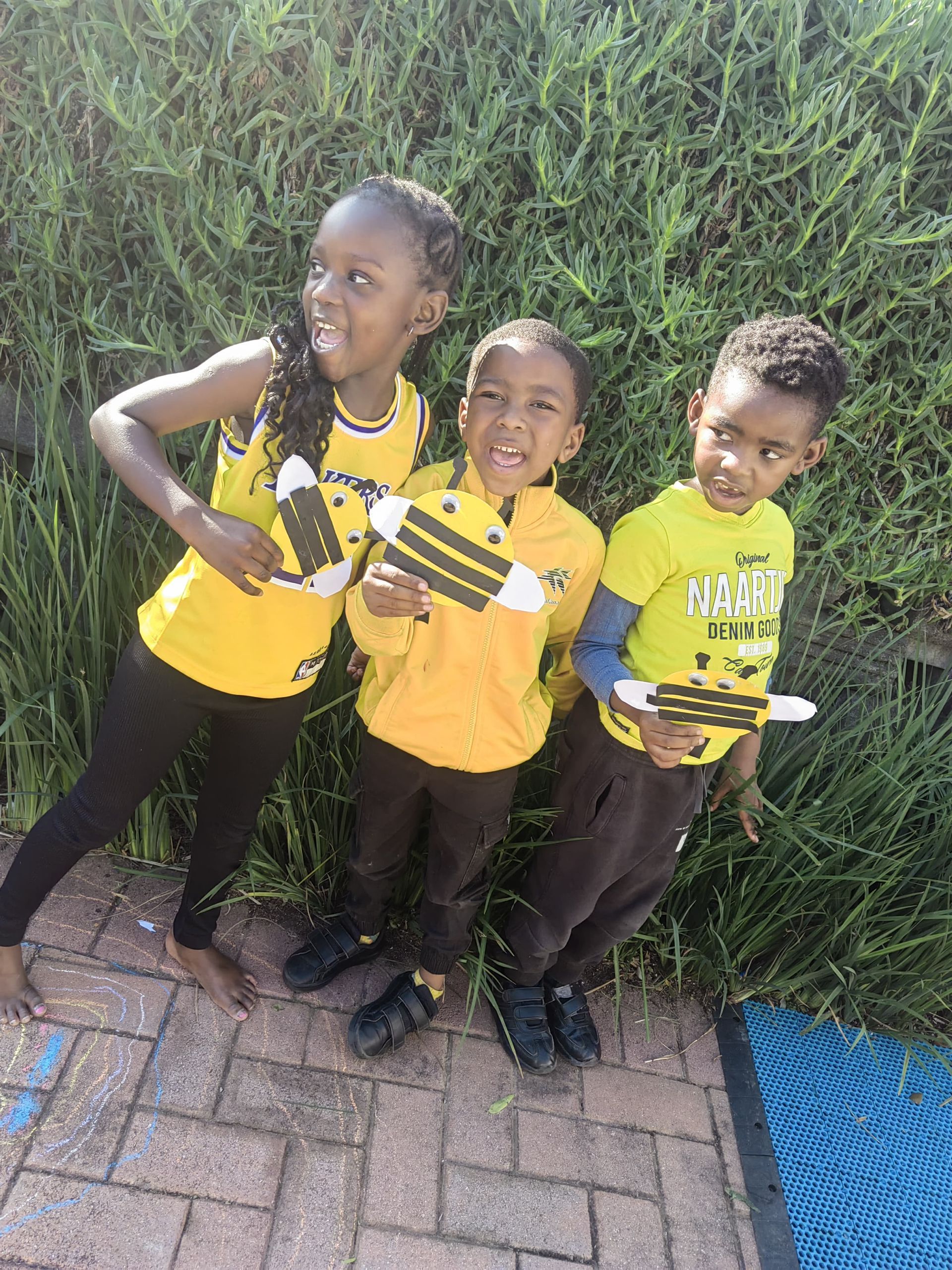 Three children in yellow holding bee-themed crafts, posing in front of greenery.