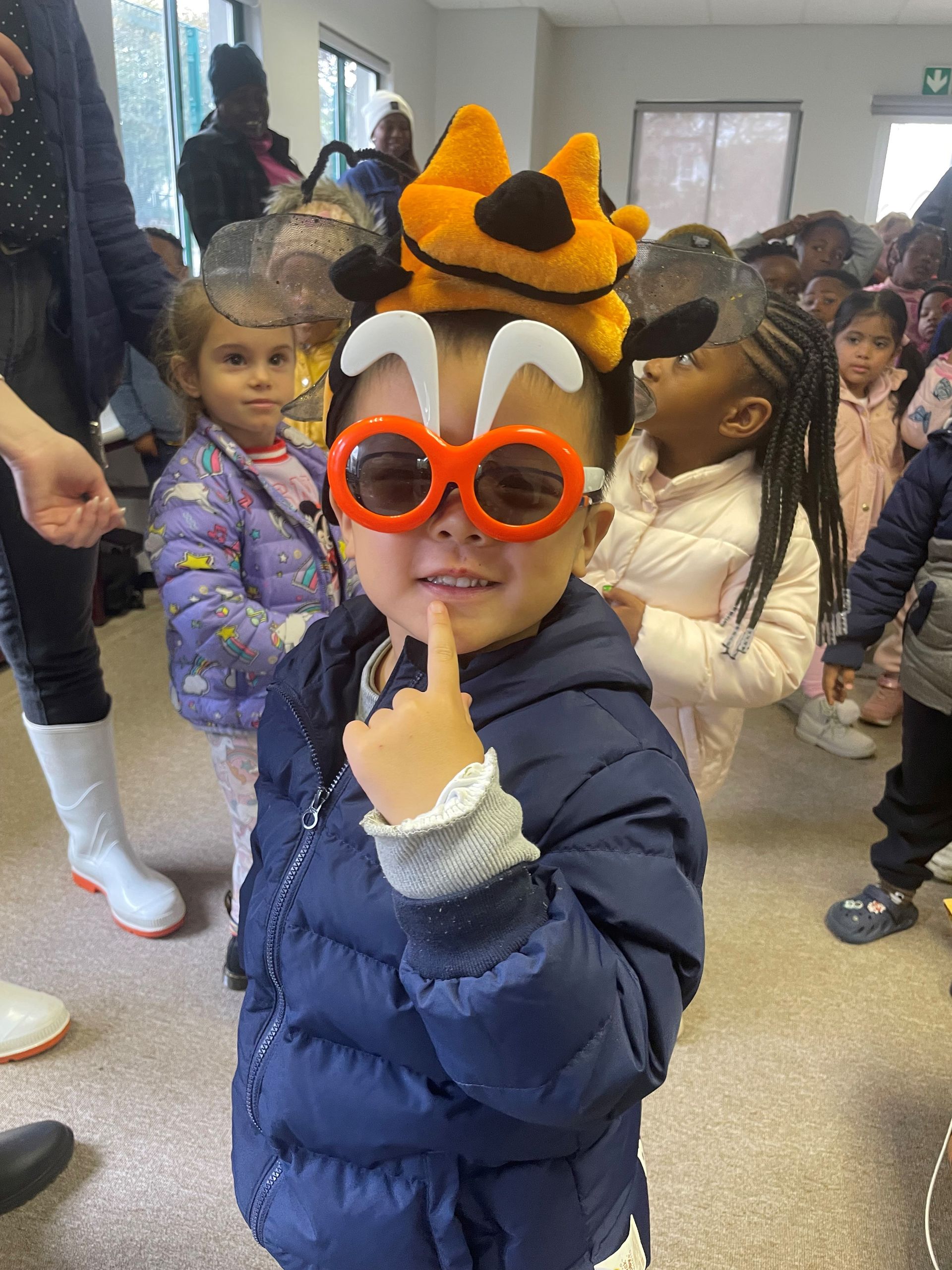 A young boy wearing a cat hat and sunglasses is standing in a room with other children.