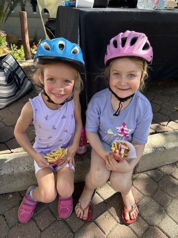 Two little girls wearing helmets are sitting on the sidewalk eating french fries.