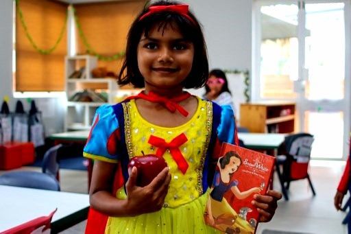 Child in Snow White costume, holding an apple and a book, smiling in a classroom.