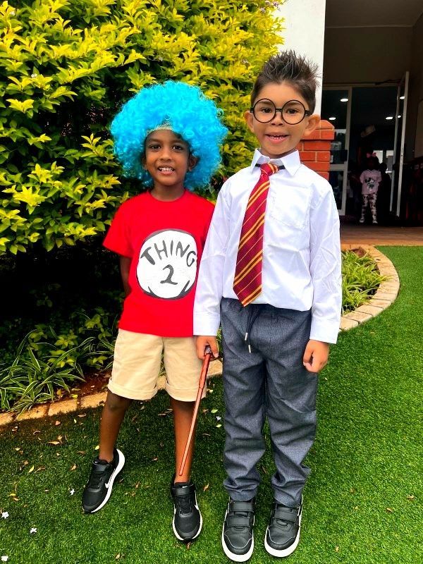 Two boys in costumes: One as Thing 2 with blue wig, the other as a professor, in front of a green bush.