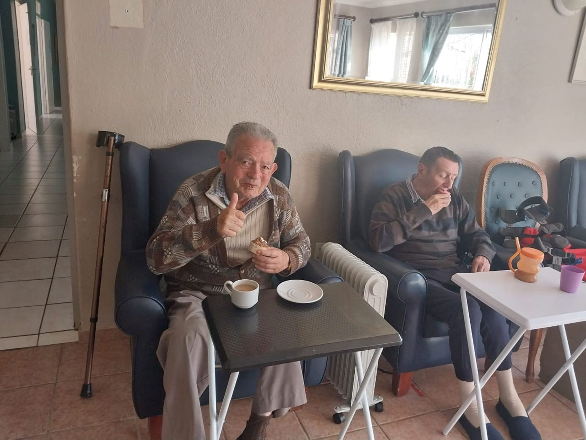 Two older men are sitting in chairs in a living room drinking coffee.