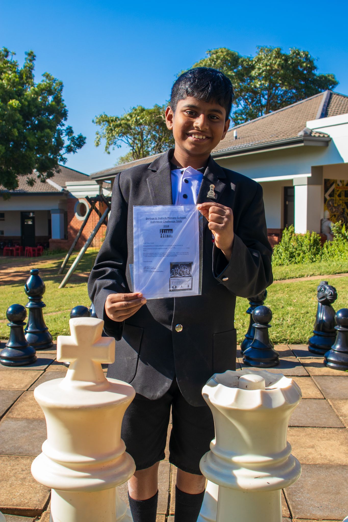 A young boy is standing next to giant chess pieces and holding a certificate.