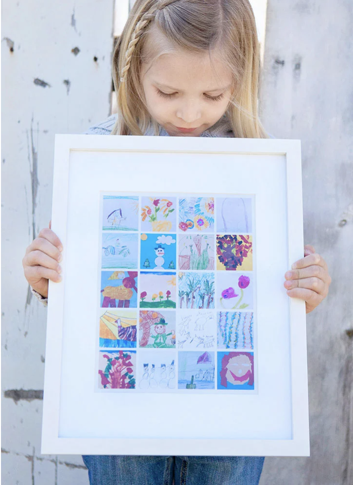 A little girl is holding a framed picture of her drawings.