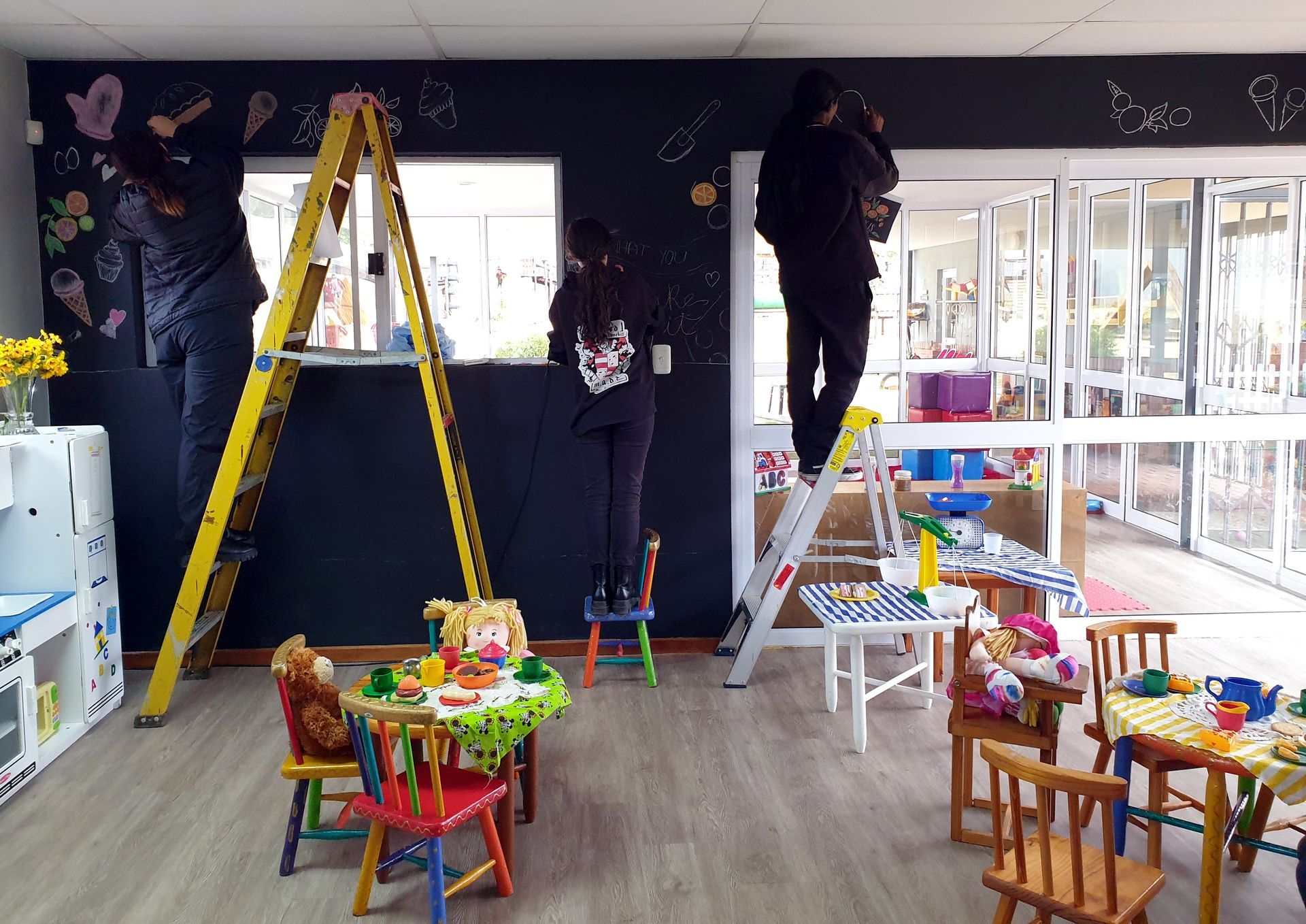 A group of people are working on a wall in a room with tables and chairs.