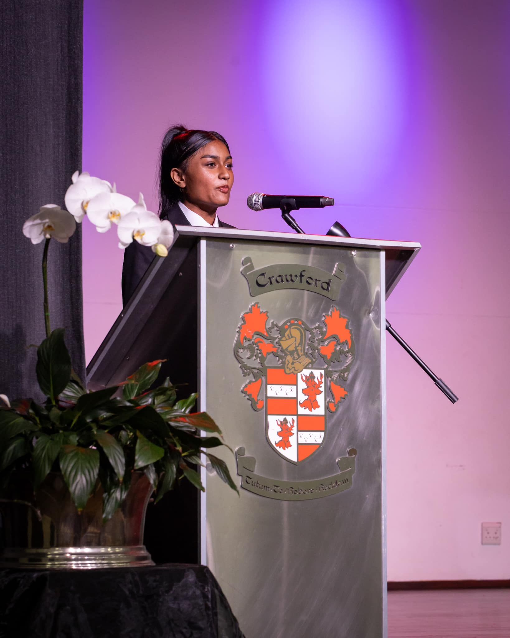 Woman speaking at a podium with a school crest. A microphone is in front of her. Flowers and plants are to her left.