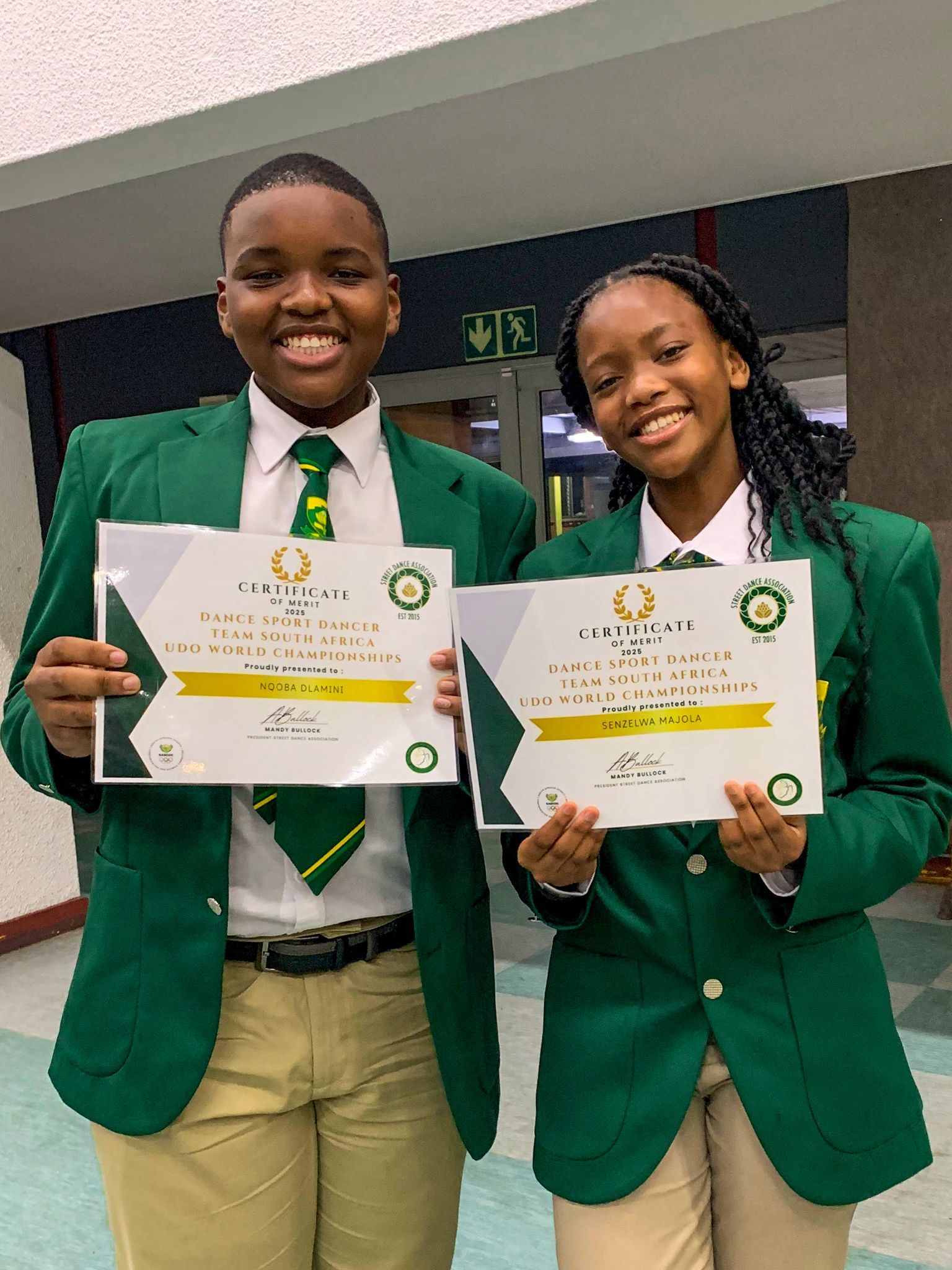 Two students in green blazers holding certificates and smiling.
