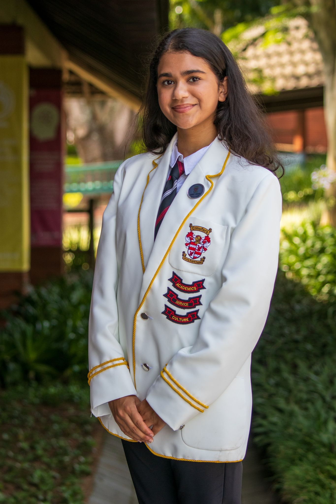 Woman in white blazer with school crest, standing outdoors, smiling.