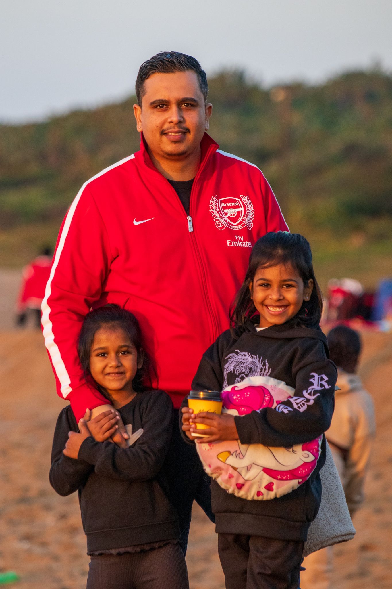 A man in a red jacket is standing next to two little girls on a beach.