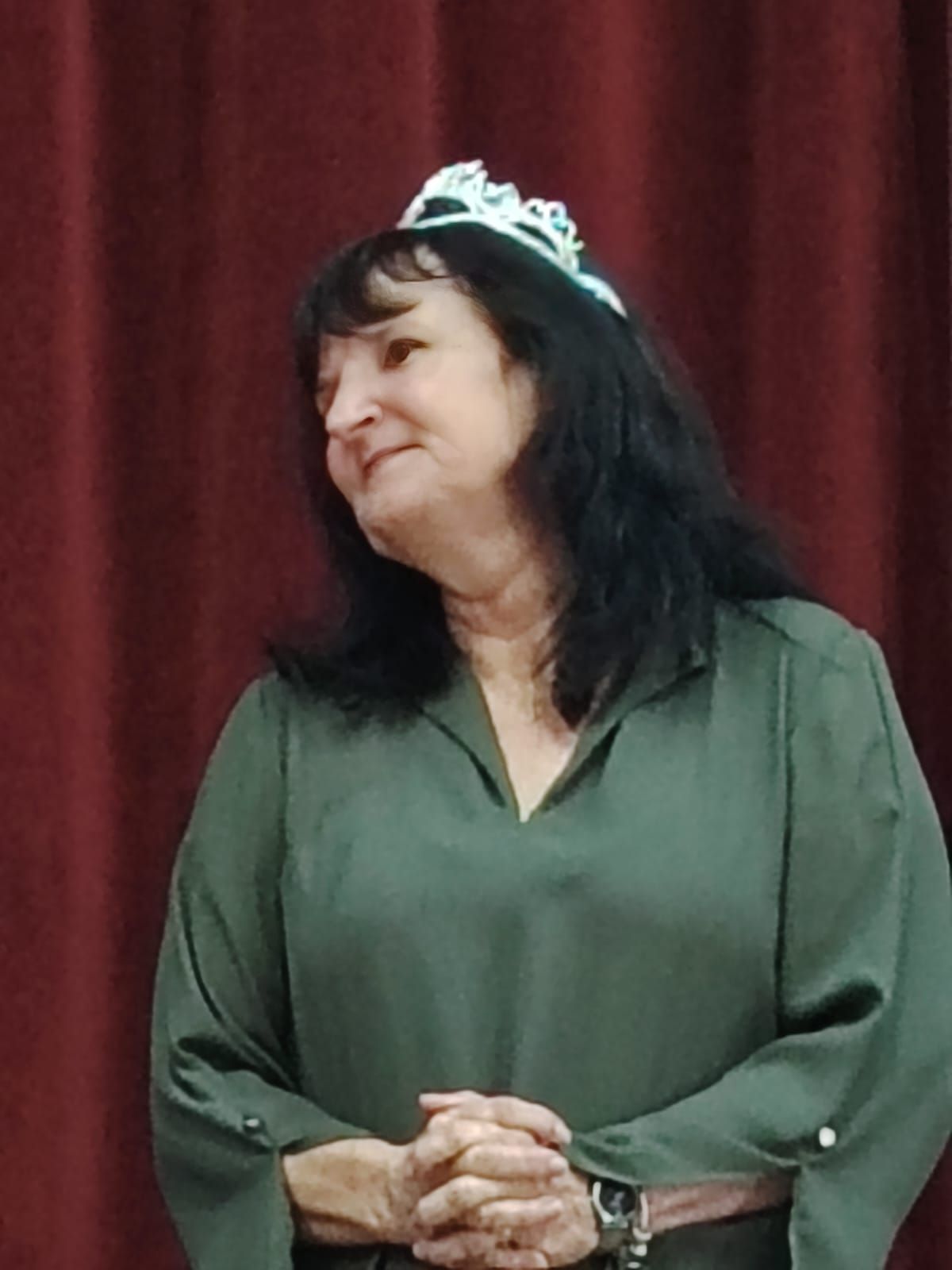 Woman with black hair, wearing a tiara and green blouse, looks to the side with hands clasped.