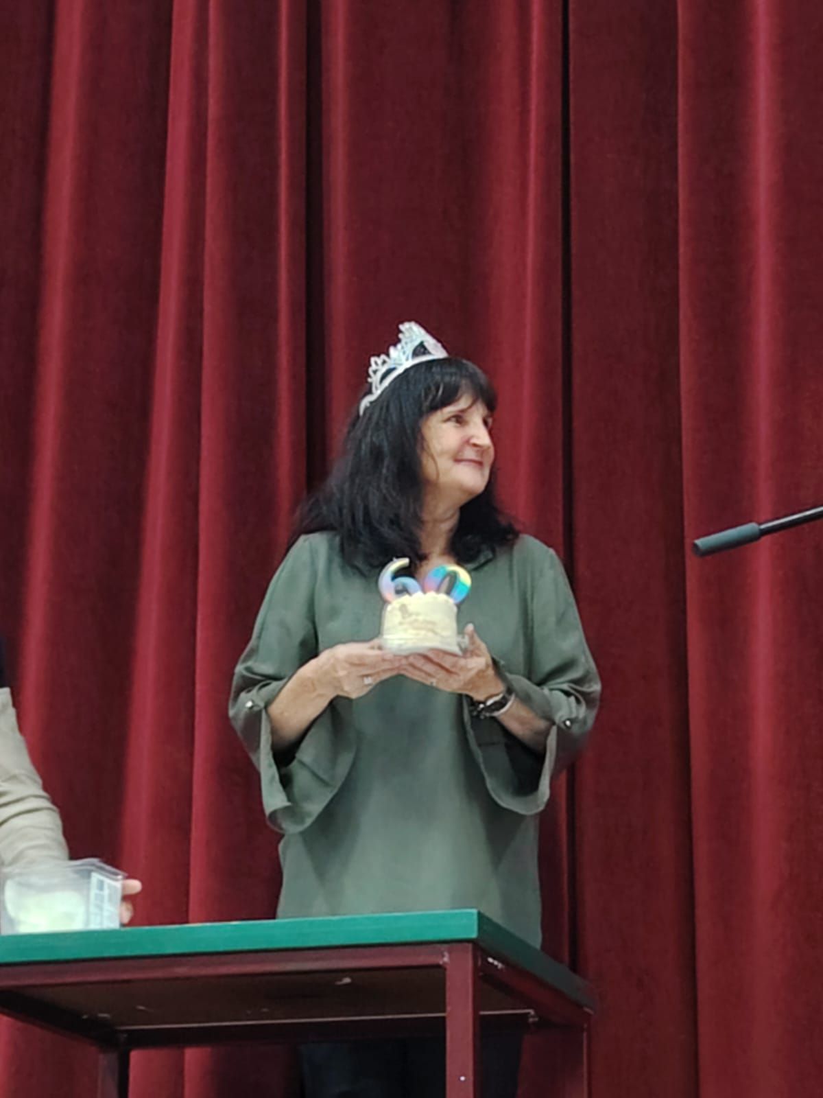 Woman in tiara holding small cake on stage with red curtains.