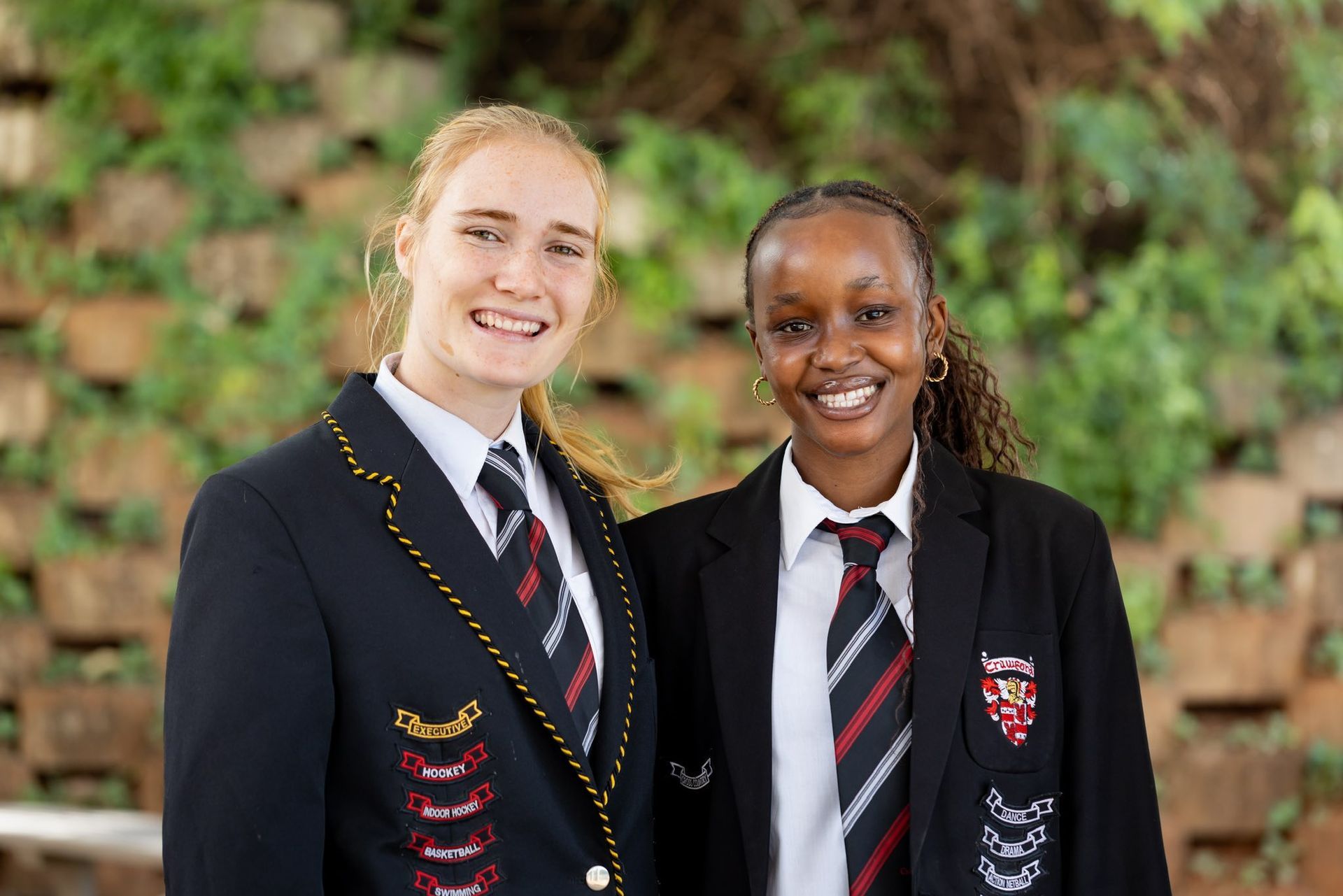 Two students in school uniforms smile, standing outside with green foliage in the background.