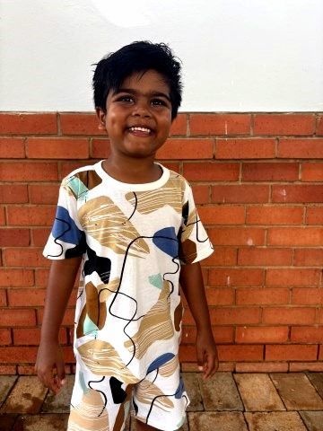 Young child smiling, standing in front of a brick wall. Wearing a white patterned shirt and shorts.