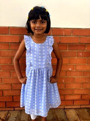Girl in blue floral dress smiles, hands on hips, against a brick wall.