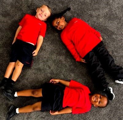 Three children in red shirts and black shorts/pants form a triangle on a gray carpet, smiling.