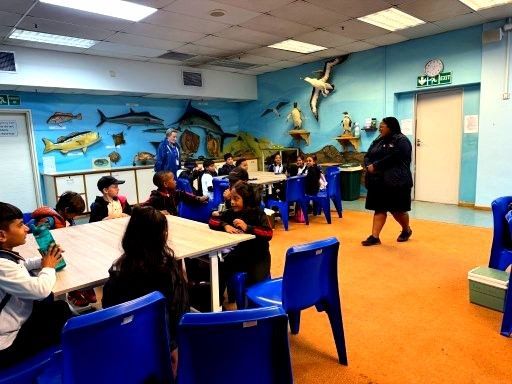 Classroom with children and an adult. Blue chairs and tables, sea life mural on the wall.