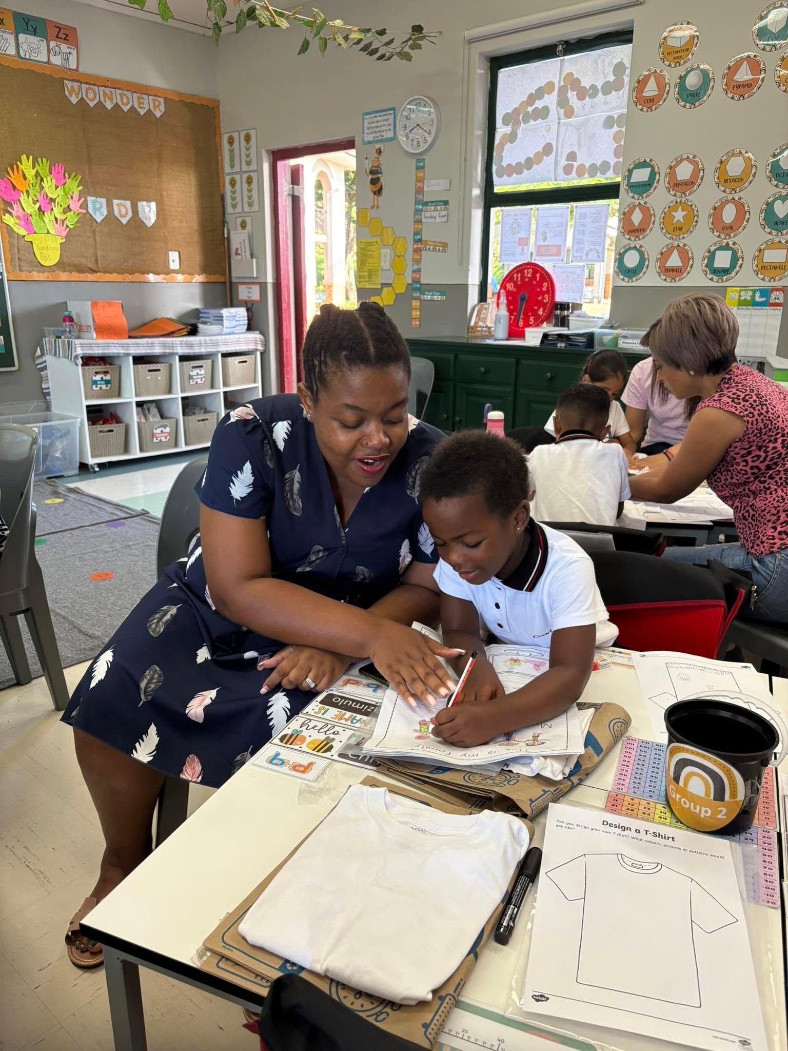 A woman helps a child with coloring at a classroom table. Other children and a teacher are in the background.