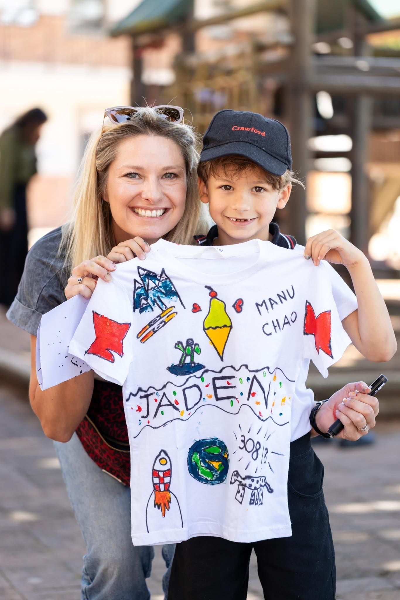 Woman and child smiling, holding a decorated white t-shirt outdoors. Shirt has colorful drawings.