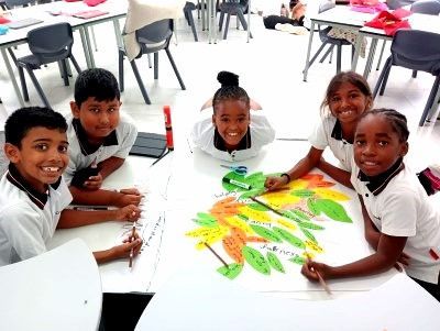 Students working on a colorful project at a white table, smiling. Bright room with desks and chairs.