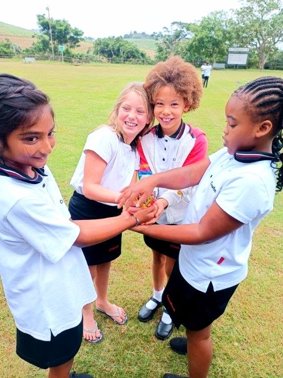 Four children in school uniforms huddle together on a grassy field, holding hands and smiling.